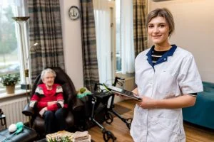 Jonge verpleegster met tablet kijkt naar camera, oudere vrouw met rode trui en looprek in huiselijke kamer