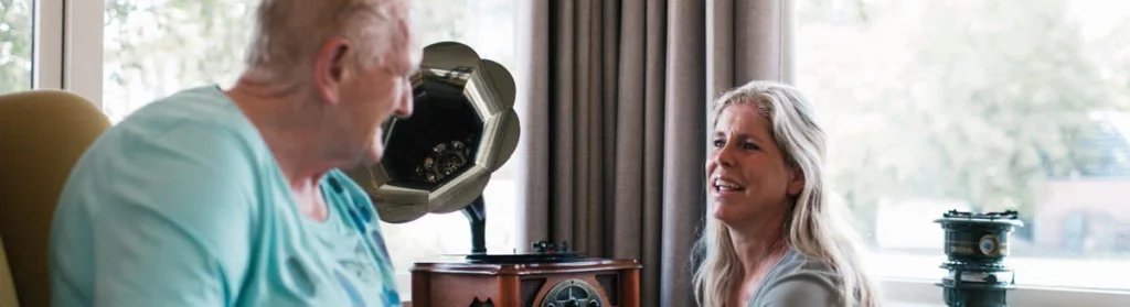 Oudere man in stoel kijkt naar vrouw die glimlachend met hem praat, grammofoon op achtergrond