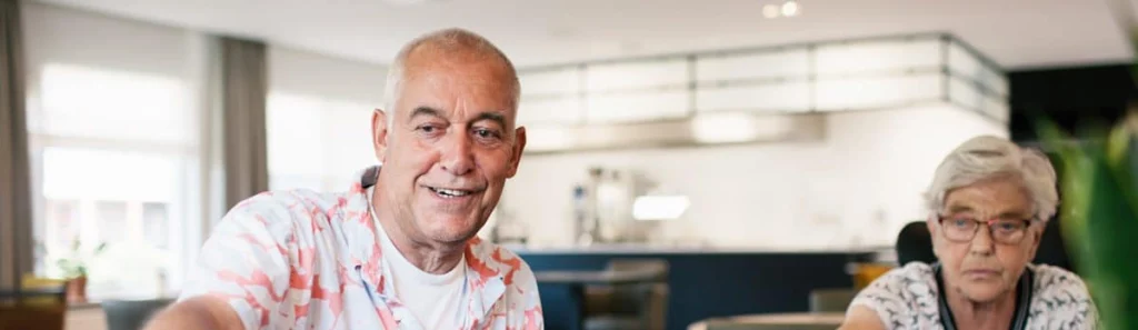 Twee oudere mensen zitten aan een tafel, de man lacht in een kleurrijk shirt, de vrouw met grijs haar draagt een bril