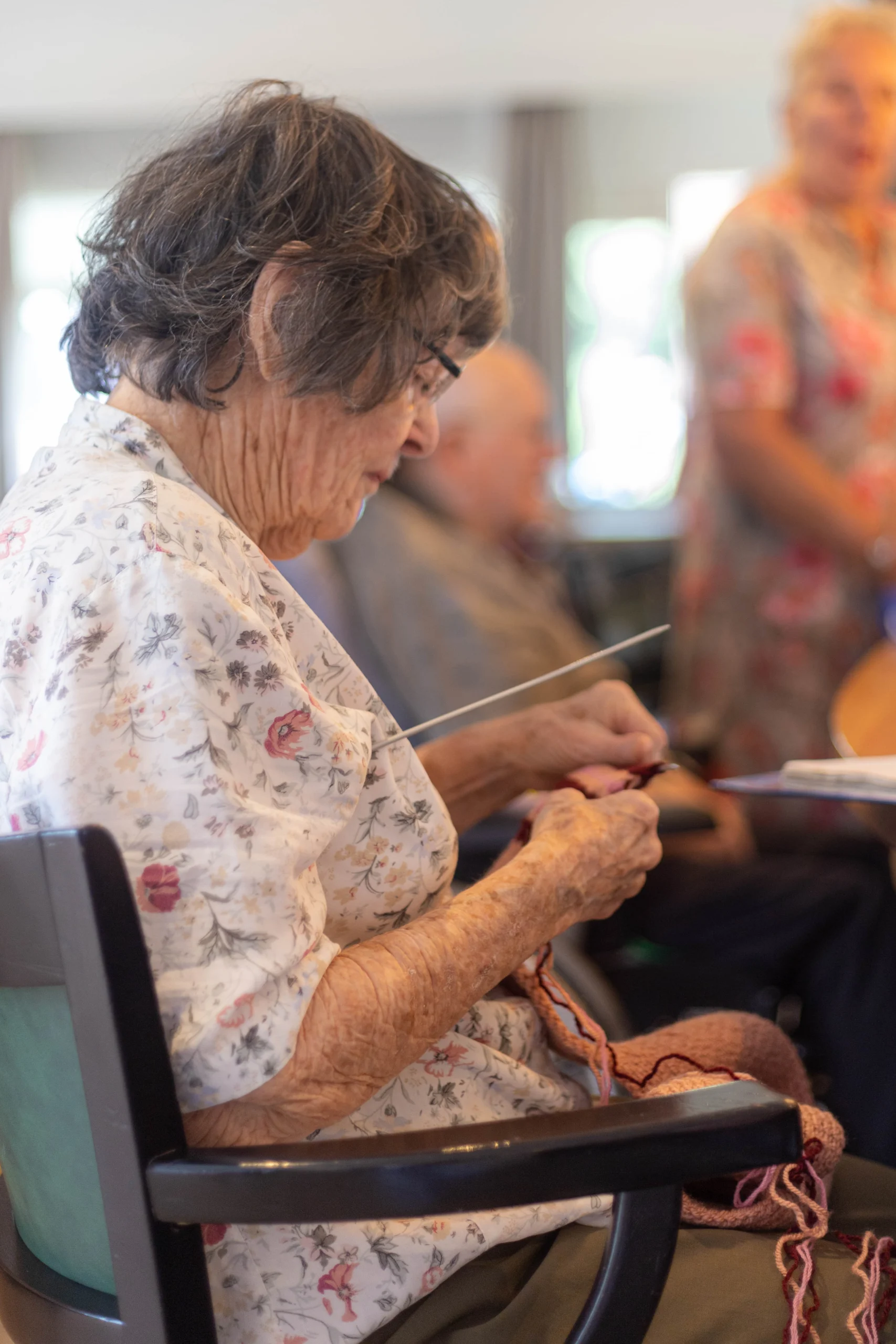 Oudere vrouw met kort krullend haar breiende aan tafel, zichtbaar breinaald en bloemmotief blouse