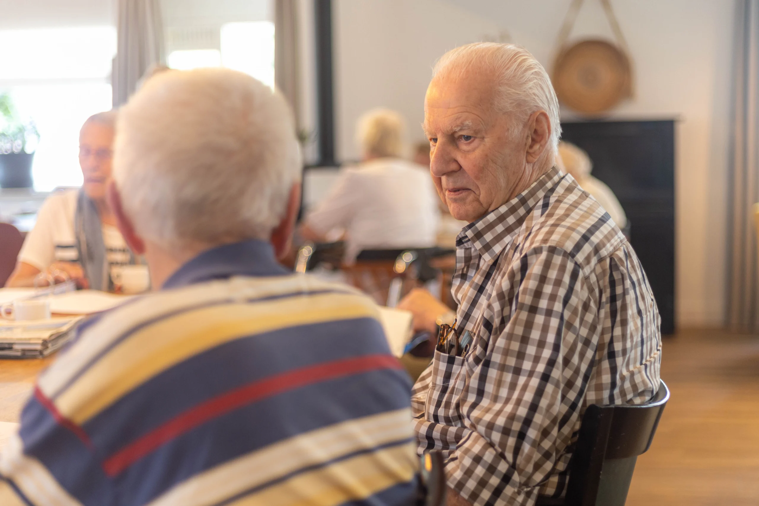 Gesprek tussen oudere mannen in een lichte, warme ruimte met natuurlijk licht en sociaal meubilair.