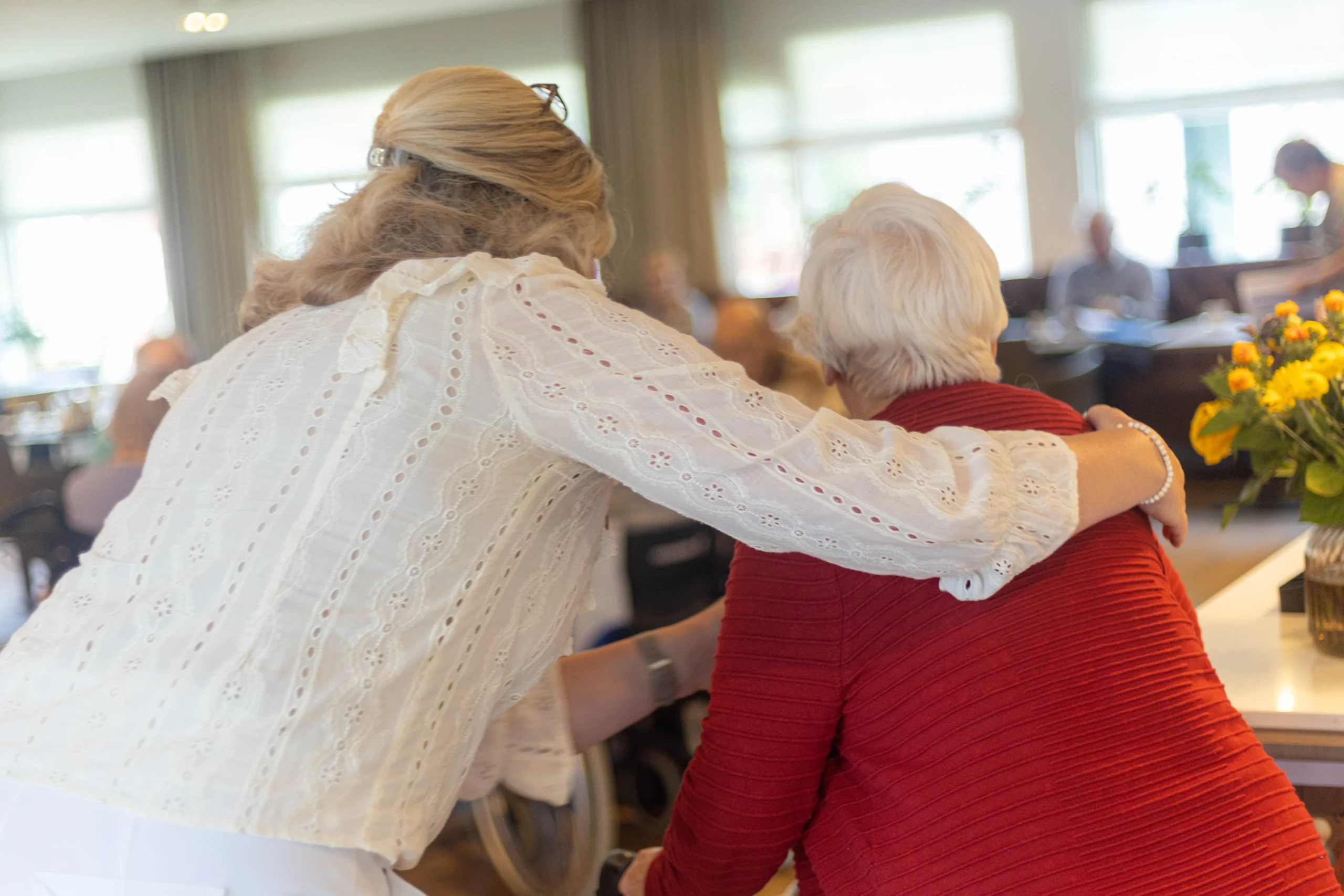 Twee vrouwen omhelzen elkaar in een gezellige ruimte met mensen op de achtergrond en een bloemstuk op tafel.