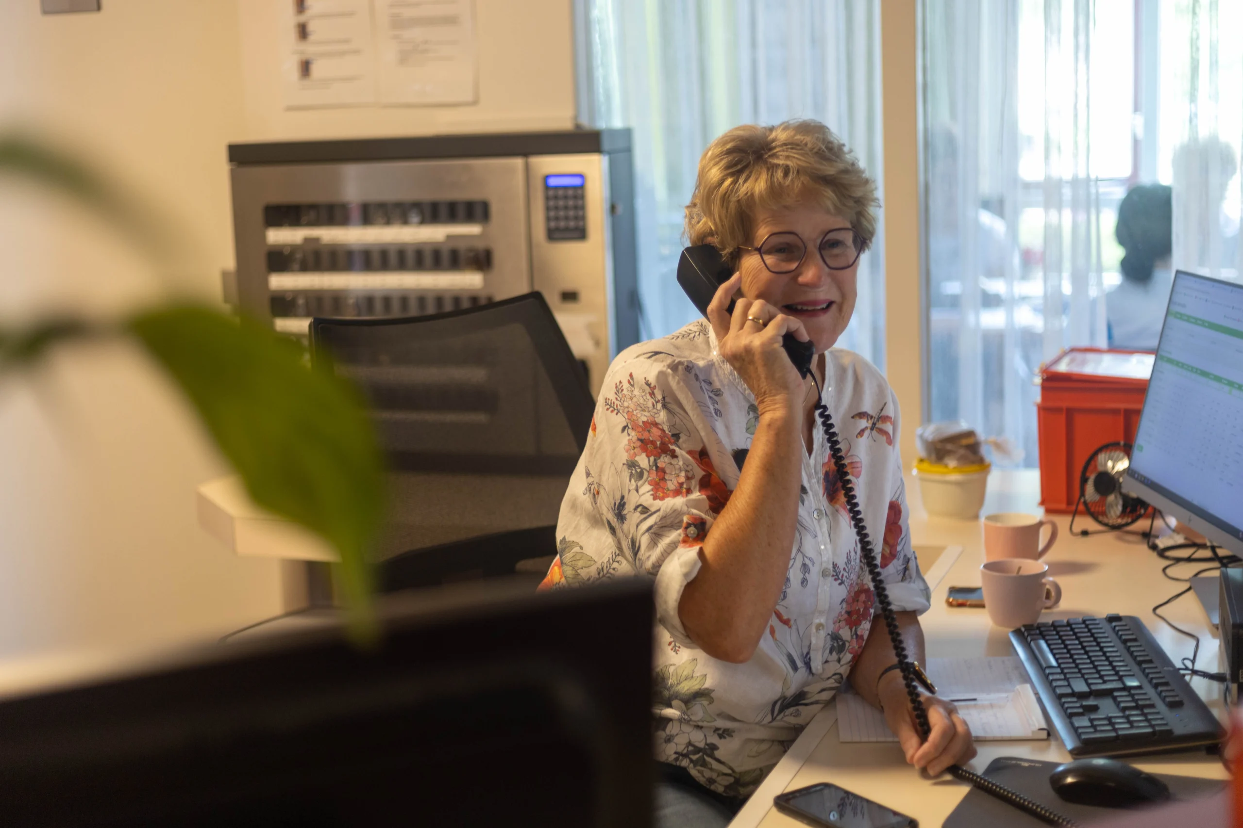 Vrouw zit aan bureau met telefoon in hand, glimlachend tijdens telefonisch gesprek, bloemenblouse aan