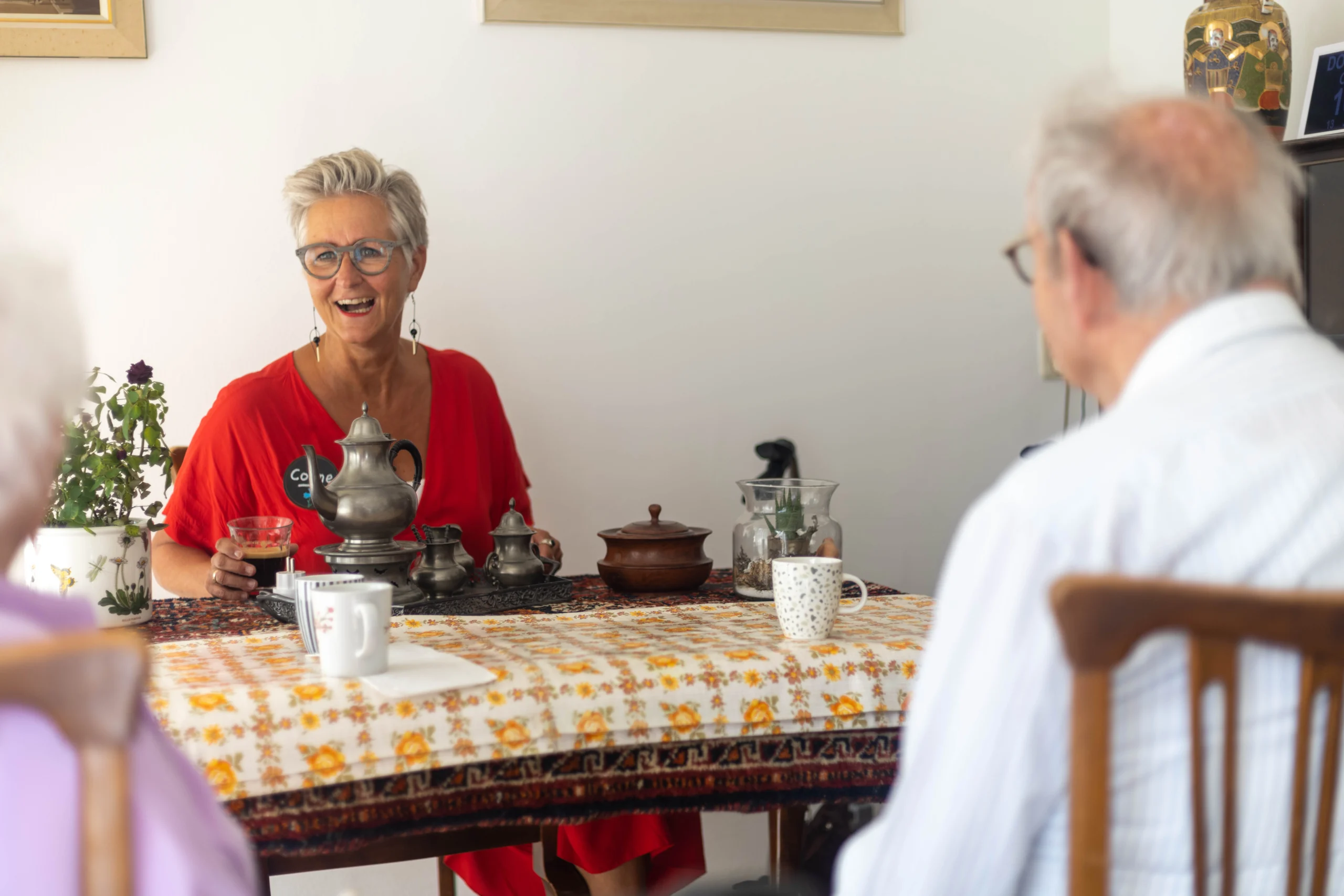 Drie personen zitten aan een gedekte tafel met theepotten en kopjes, vrouw in rode outfit lacht