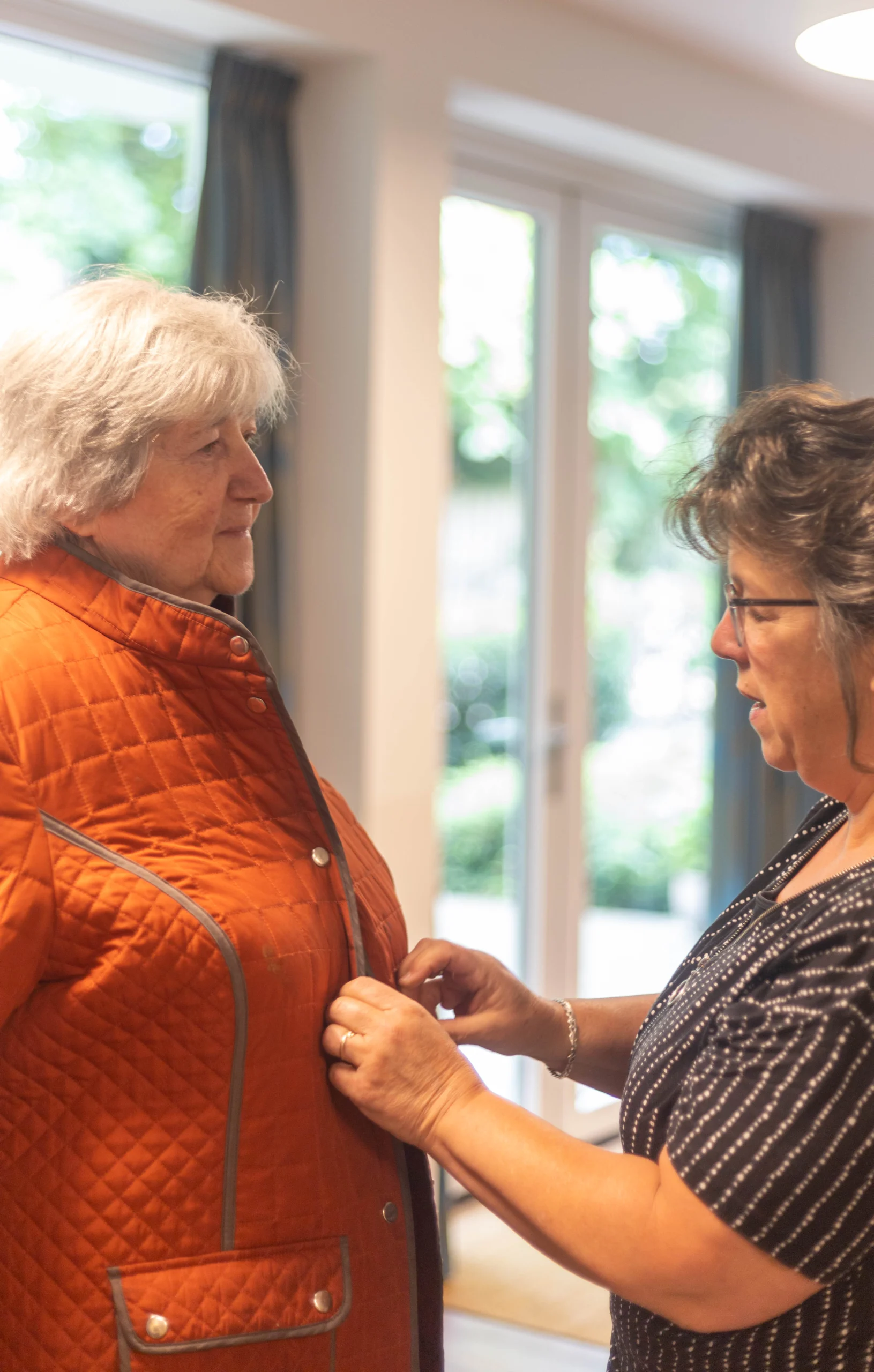 Twee vrouwen in een lichte binnenruimte waarbij de rechter vrouw de jas van de linker vrouw dichtknopt.