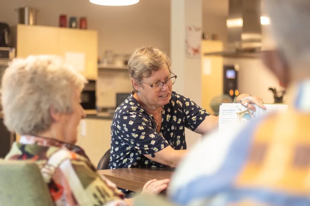 Een vrouw met grijzend kort haar in een blauwe blouse wijst iets aan in een lichte woonkamer met ouderen