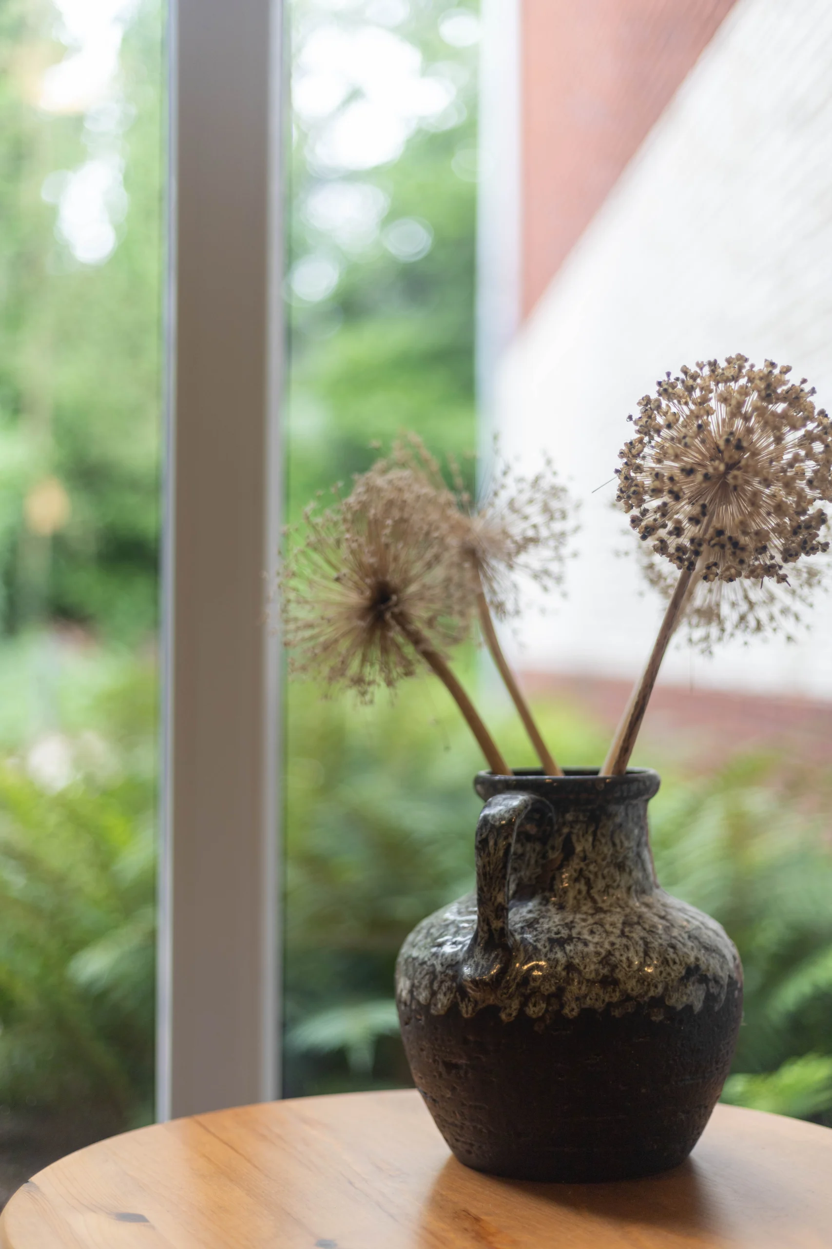 Zwart aardewerken vaasje met handvat op houten tafel met drie gedroogde bloemen en raam op achtergrond