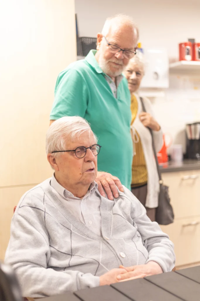Drie ouderen in een warme gemeenschappelijke ruimte met keukenachtergrond, in gesprek en ontspannen.