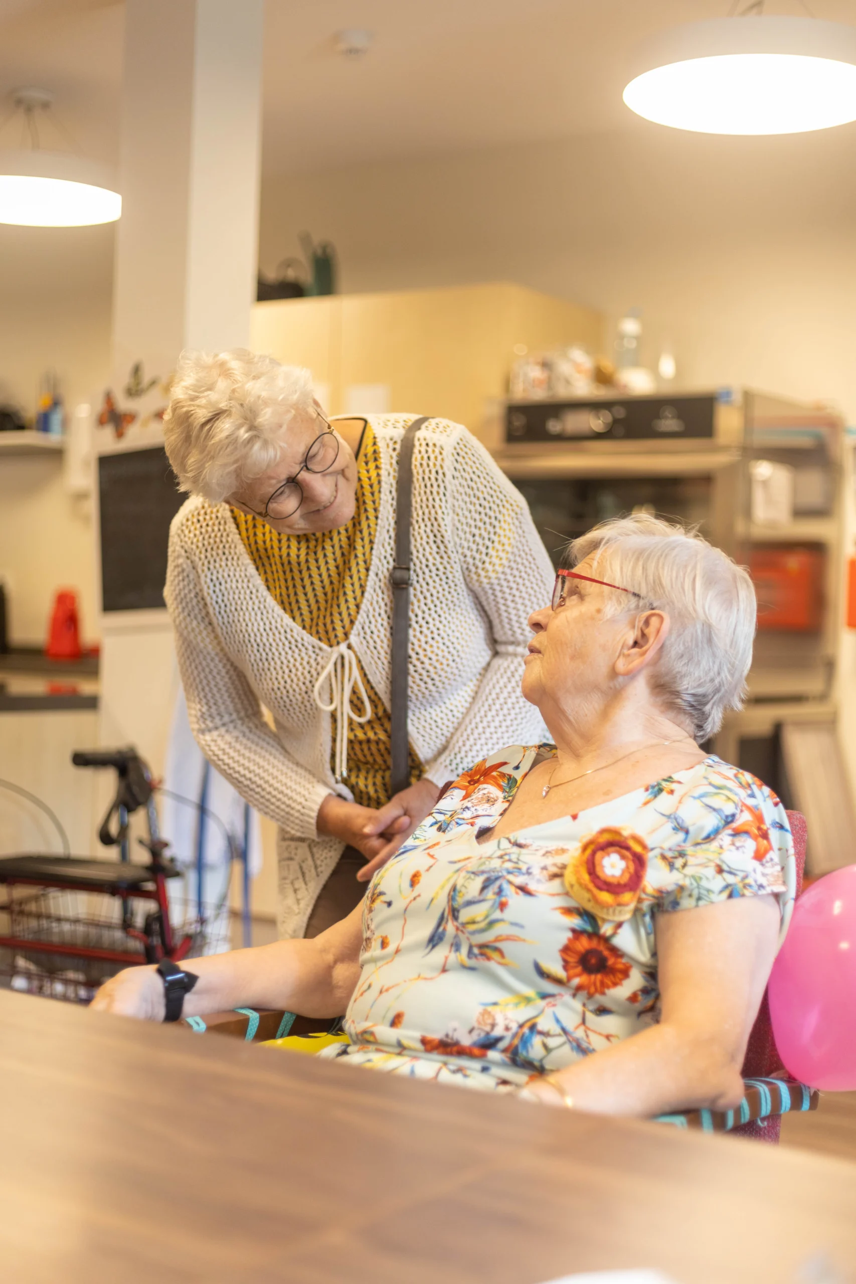 Twee oudere vrouwen praten ontspannen in een keukenachtige ruimte met ballonnen en een rolstoel op de achtergrond.