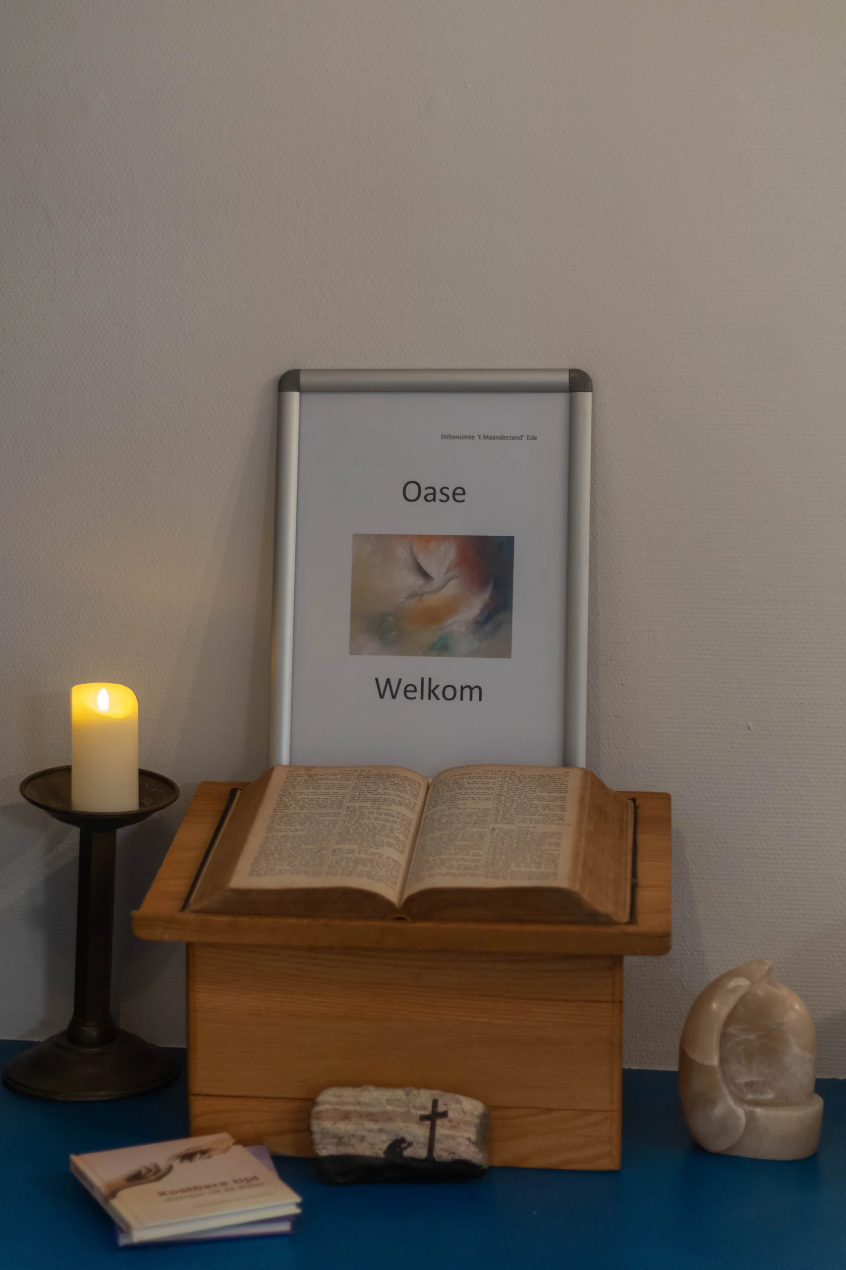 Houten boekenkast met een open oud boek, kaars, stenen kruis en bord met tekst 'Oase Welkom'