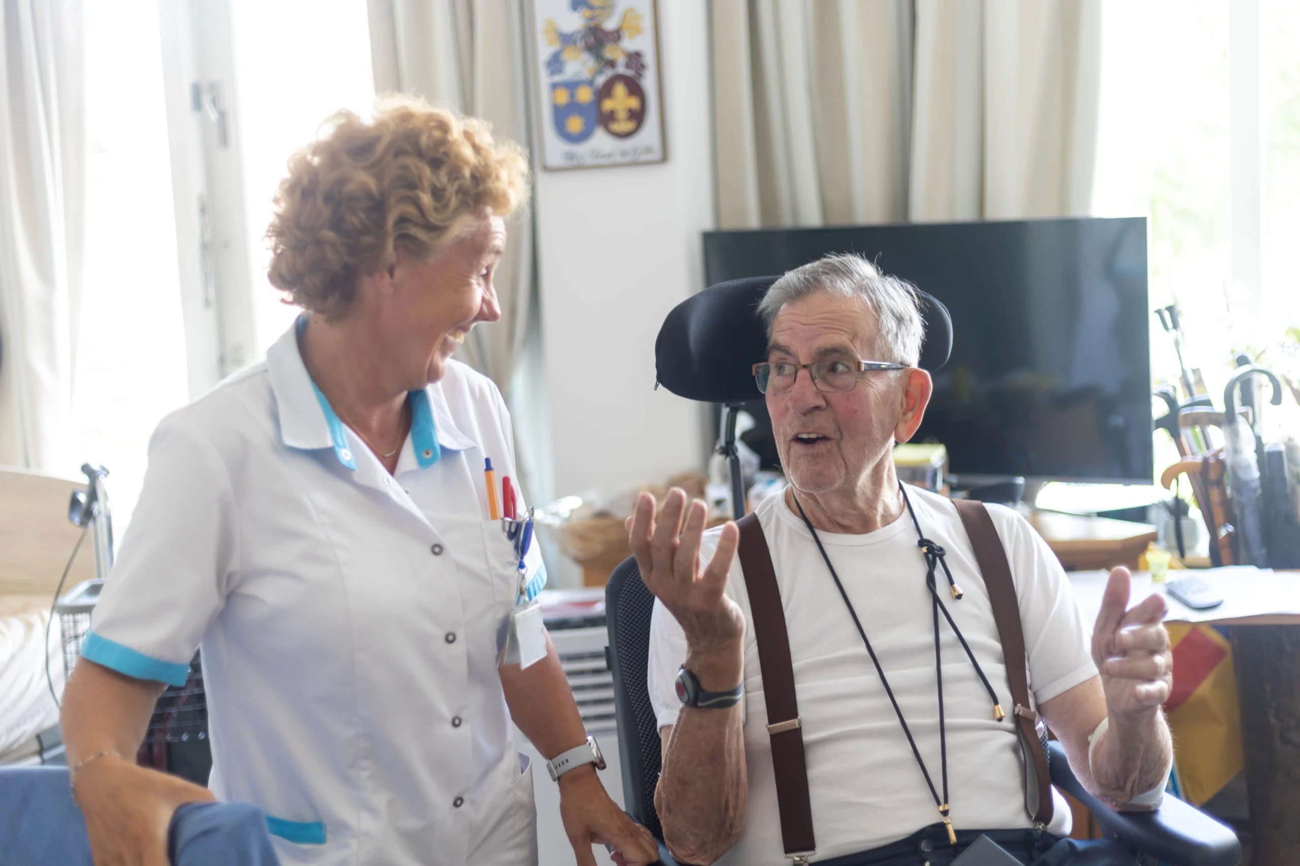 Zorgverlener in witte labjas praat lachend met oudere man in rolstoel in huiselijke kamer