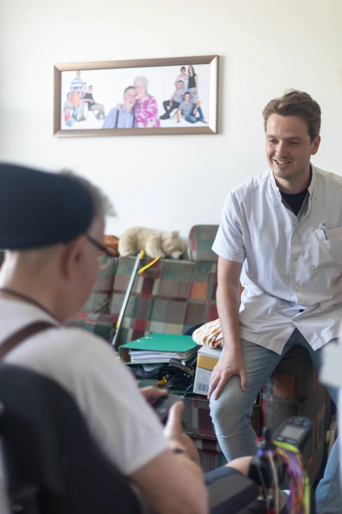 Gesprek tussen jongeman in lichte blouse en oudere man in rolstoel in huiselijke woonkamer.