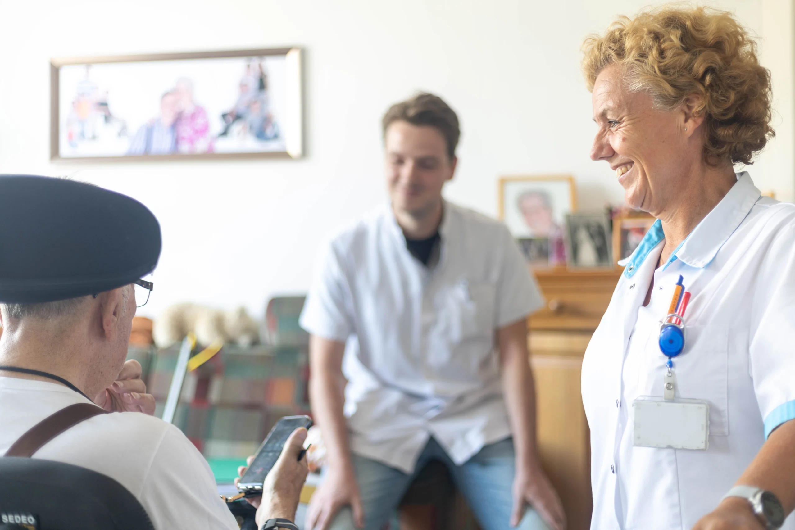 Drie mensen in een huiselijke ruimte waarbij een oudere man met zwarte hoed op zijn smartphone kijkt