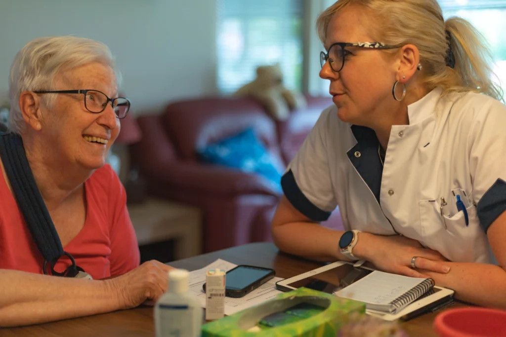 Een oudere vrouw met bril lacht naar een zorgverlener aan tafel met smartphone en papieren.