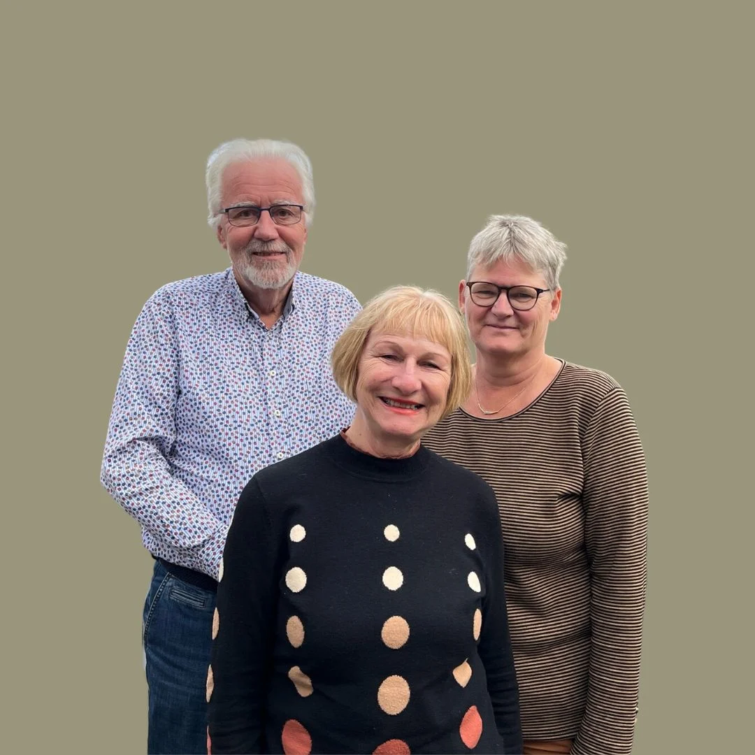 Drie volwassenen tegen een beige achtergrond, man met grijs haar en baard achter twee glimlachende vrouwen.