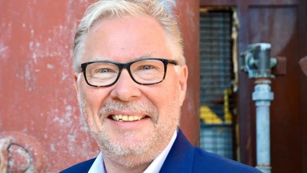 Man met grijs haar en lichte baard draagt brede bril en blauwe blazer buiten bij gekleurde muur