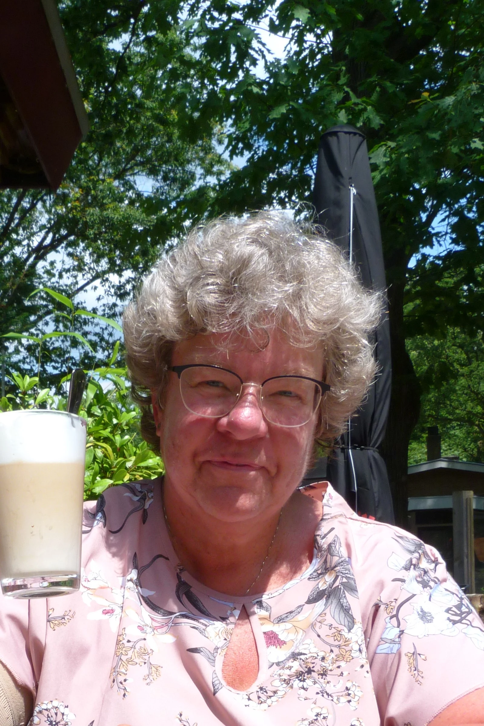 Vrouw met gekrulde haren en lichte bloes met bloemen houdt glas met romig drankje buiten vast.