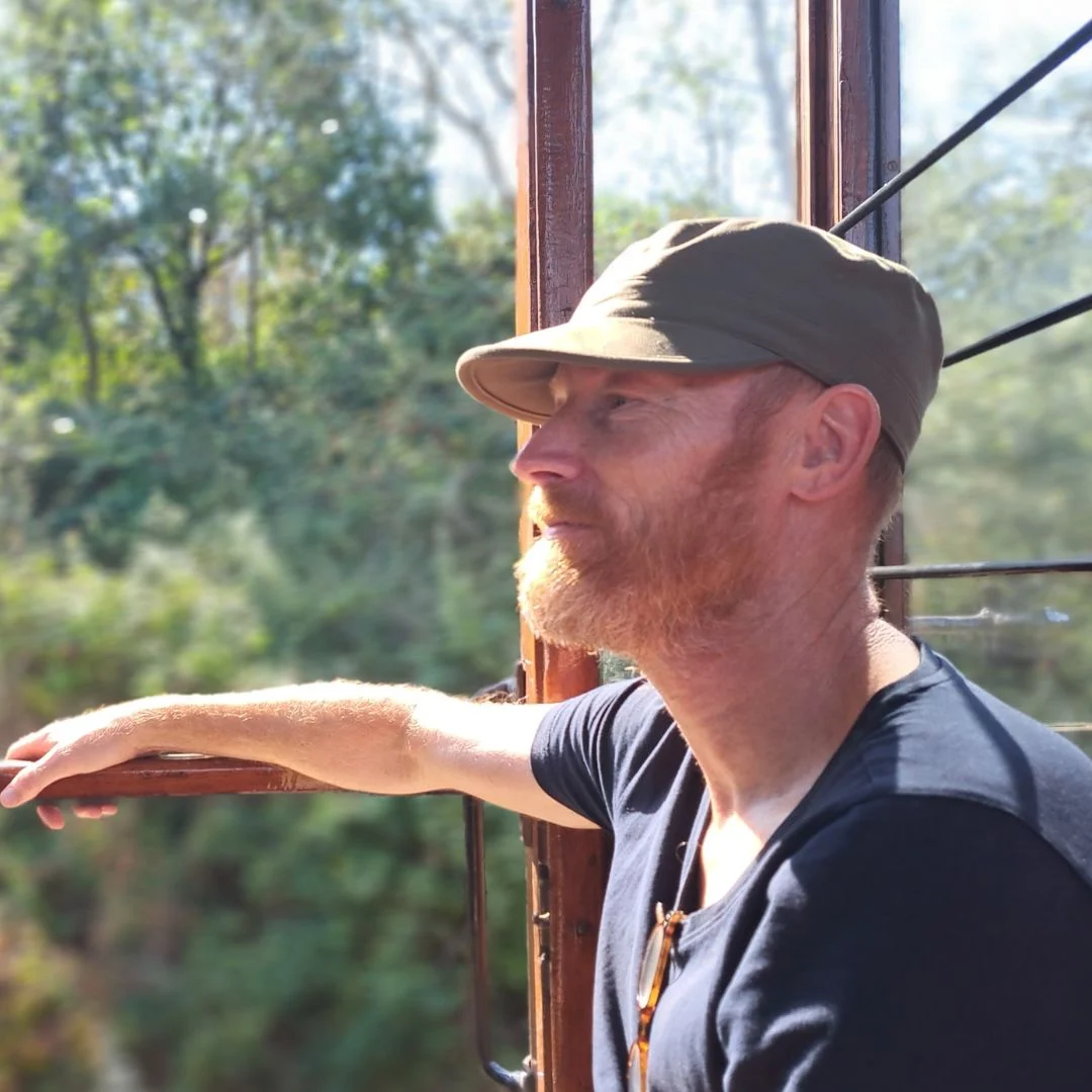 Man met kort rossig baard en groene pet zit ontspannen buiten in een natuuromgeving bij houten rand.