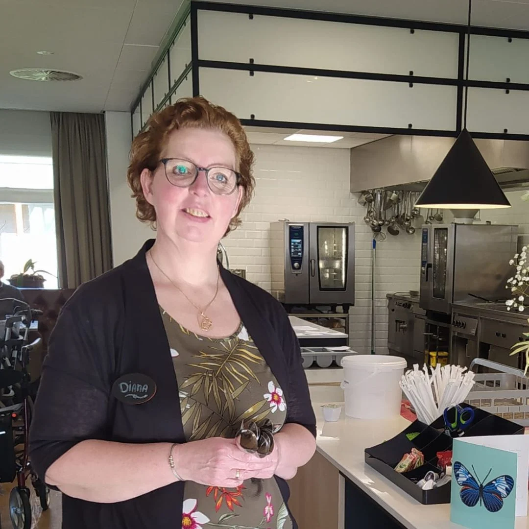Vrouw met kort krullend haar houdt zonnebrillen vast in een moderne keuken met keukentoestellen en rolstoel.