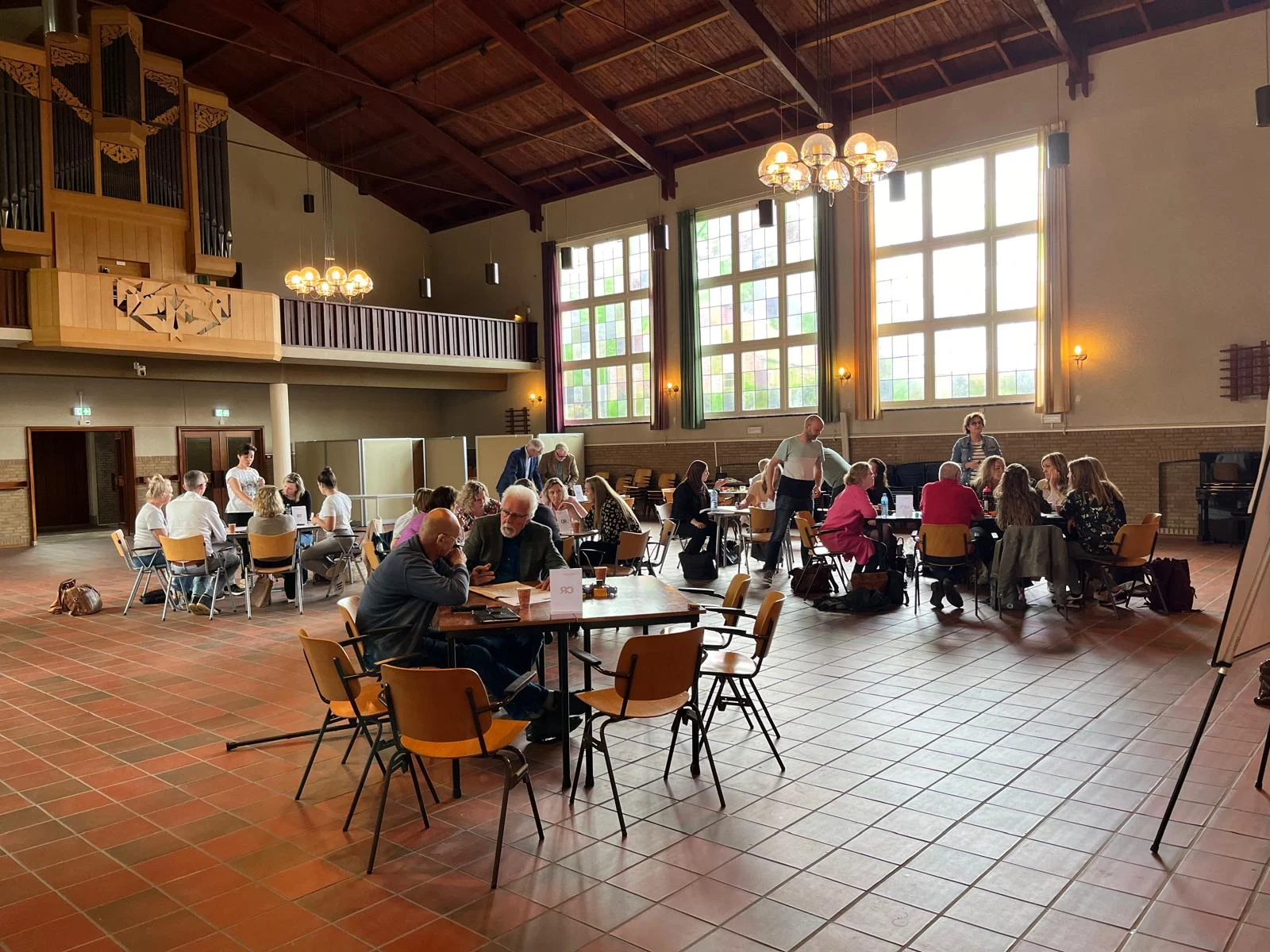 Grote lichte zaal met hoog houten plafond, orgel aan muur en mensen die aan tafels samenwerken.