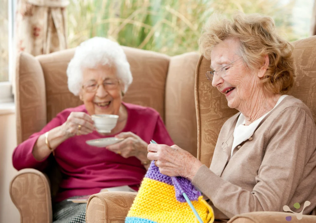 Twee oudere vrouwen zitten in comfortabele stoelen, één drinkt thee, de ander breit lachend
